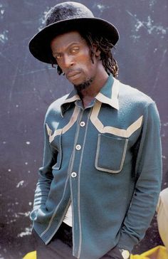 a man with dreadlocks wearing a green shirt and black hat standing in front of a wall