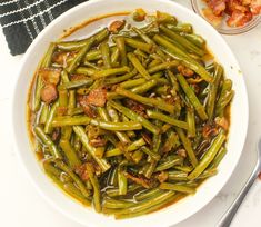 a white bowl filled with green beans next to some other food on a table and utensils