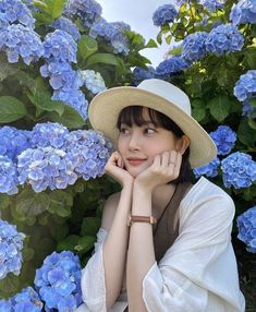 a woman wearing a hat sitting in front of blue hydrangea flowers with her hands on her chin