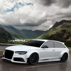 a white car driving down a road with mountains in the background and cloudy skies above