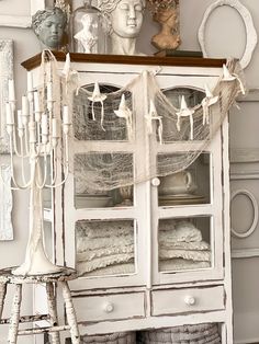 an old white china cabinet with glass doors and curtains on the top, next to a stool