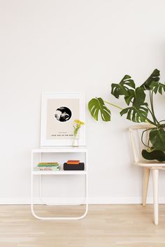 a white shelf with books and plants on it in front of a wall mounted poster