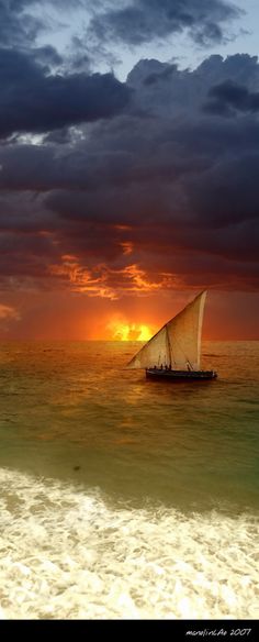 a sailboat in the ocean at sunset with dark clouds above it and an orange sky