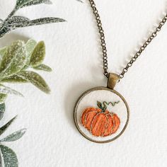 an embroidered pumpkin is hanging from a chain on a white surface with green leaves in the background