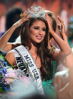 a beautiful woman wearing a tiara and holding her hands up to her head while standing in front of a crowd