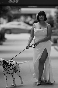 a woman in a white dress walking a dalmatian on a leash down the street