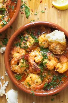 two bowls filled with shrimp and sauce on top of a wooden table next to lemon wedges