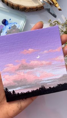 a person holding up a small painting in their hand with paintbrushes on the table