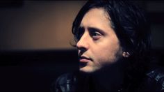 a man with long black hair wearing a leather jacket and looking off into the distance