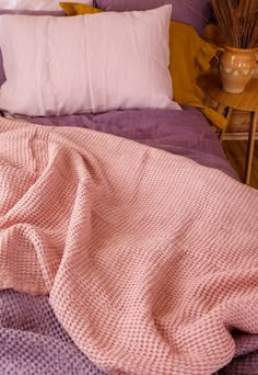 an unmade bed with pink and purple blankets on it, next to a potted plant