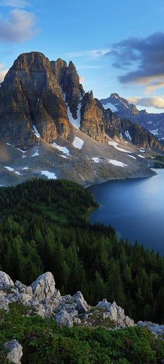 the mountains are covered in snow and green trees, with blue water below them on a sunny day