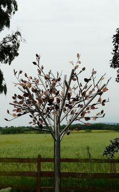 a metal tree with leaves on it in the middle of a grassy field next to a wooden fence