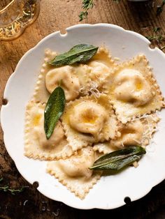 a white plate topped with ravioli covered in cheese and sage leaves next to a glass of wine