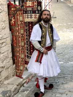 a man dressed in traditional clothing standing next to some rugs on the street and looking at the camera