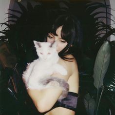 a woman holding a white cat in her arms while sitting on a chair next to plants