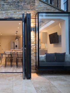 an open glass door leading into a living room and dining area with brick walls, floor to ceiling windows, and outdoor furniture