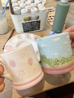 someone is holding two coffee mugs in front of some crafting supplies on a table