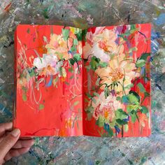 a hand holding an open book with flowers painted on the cover and in front of it