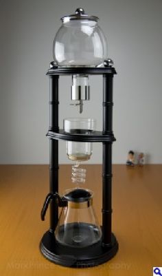 a person pouring water into a glass coffee pot on top of a wooden table next to an espresso machine