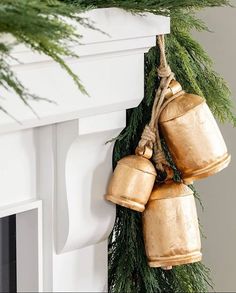 two golden bells hanging from a green garland on a fireplace mantel with pine branches