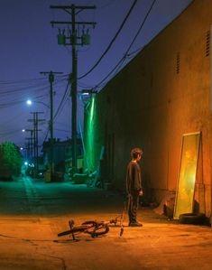 a man standing in the middle of an empty street at night with his bicycle leaning against a wall