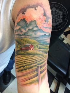 a man's arm with a farm scene on it and clouds in the sky