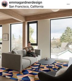 a person sitting on a couch in a room with large windows and colorful rugs