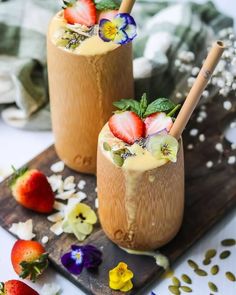 two drinks with strawberries and flowers on a wooden cutting board next to other food items