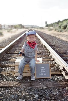 a young boy sitting on top of a wooden crate next to train tracks with a chalkboard