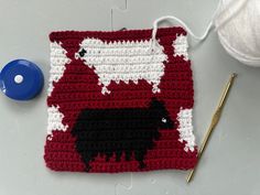 a red and white crocheted square with black sheep on it next to a ball of yarn