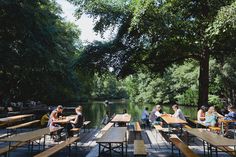 many people are sitting at picnic tables on the water's edge, with trees in the background