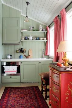 a kitchen with green cabinets, red rug and pink curtains on the window sill