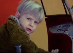 a young boy with blonde hair sitting in front of a red chair and looking at the camera