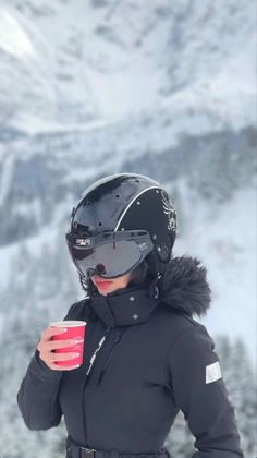 a woman wearing a helmet and holding a red cup in her hand while standing on top of a snow covered mountain