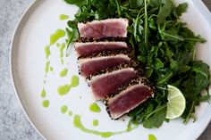 a white plate topped with slices of tuna next to greens and lemon wedges on top of a table