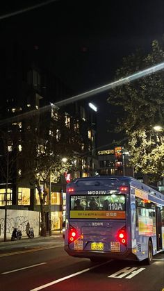 a city bus driving down the street at night with its lights on and buildings in the background