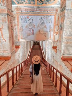 a woman in a white dress and hat walking down a flight of stairs with egyptian paintings on the walls