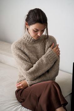 a woman sitting on top of a white couch wearing a sweater and brown skirt with her arms crossed