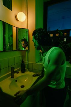 a man standing in front of a bathroom sink with green light on the mirror above it