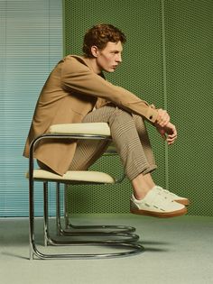 a man sitting on top of a chair in front of a green wall and wearing a tan coat