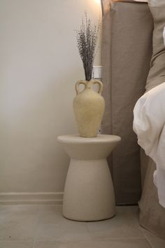 a white vase sitting on top of a small table next to a bed with pillows