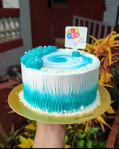 a hand holding a cake with blue icing and white frosting in front of flowers