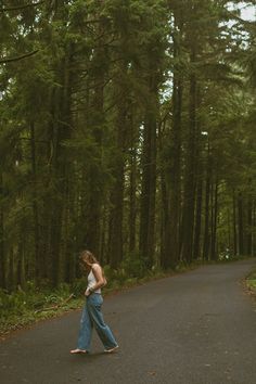 a woman walking down a road in the middle of a forest with tall trees on both sides