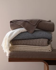 a stack of folded sweaters and blankets on top of a wooden table next to a wall