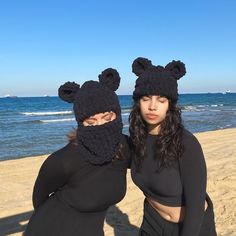 two women in black outfits standing on the beach with their faces covered by bear ears