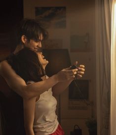 a man and woman standing next to each other in front of a window looking at their cell phones