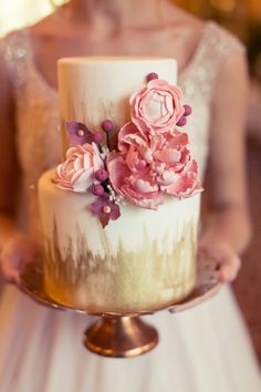 a white and gold wedding cake with pink flowers on the top is being held by a woman