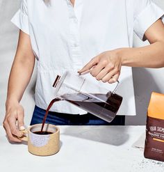 a woman pours coffee into a cup from a pitcher, with two bags of ground coffee nearby