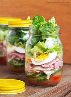 three mason jars filled with salads on top of a wooden table next to yellow lids