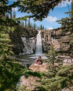 two people sitting on the edge of a cliff looking at a waterfall in the distance